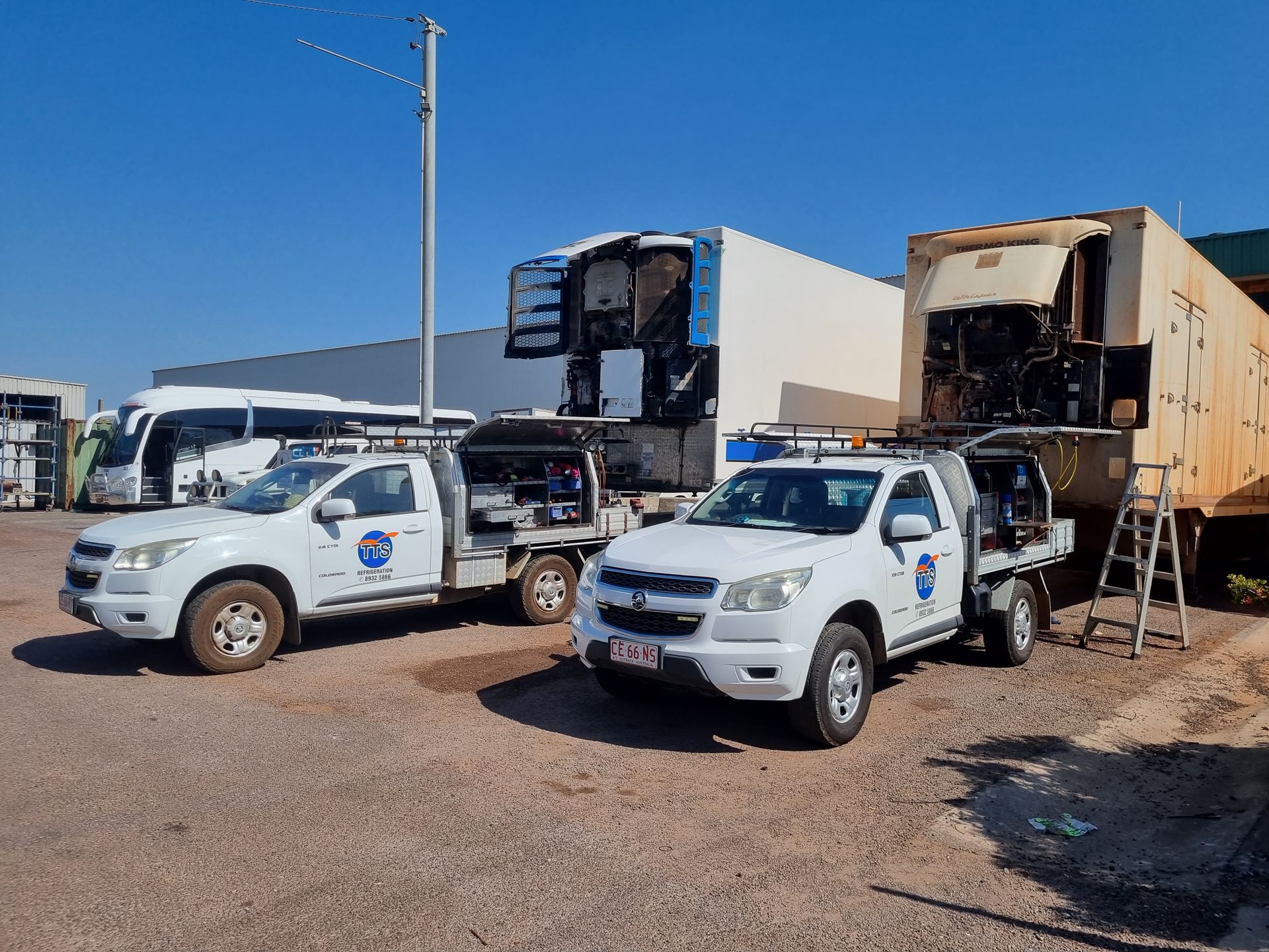 Two white pickup trucks with equipment next to large industrial vehicles in a paved area under a blue sky — TTS Refrigeration in Pinelands, NT