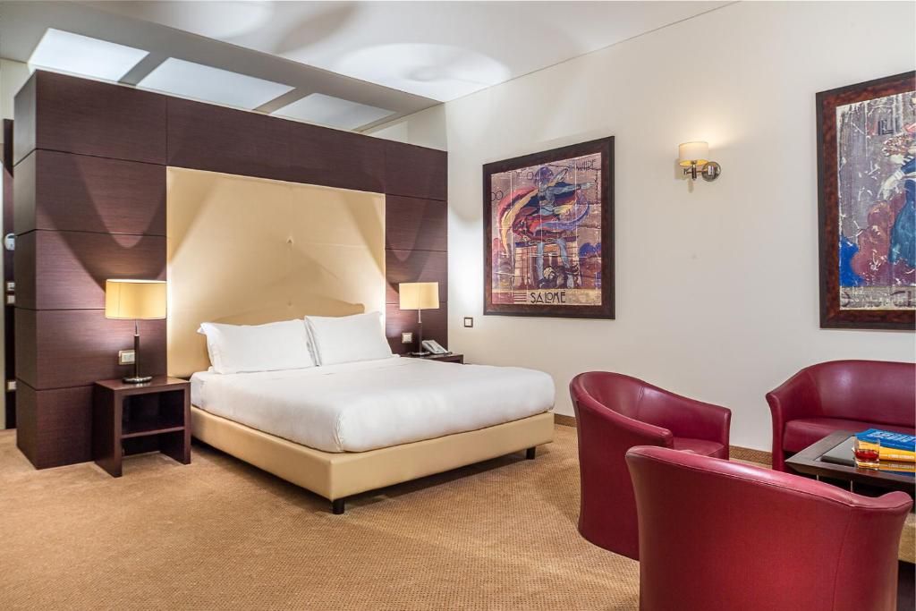 Camera d'albergo moderna con letto king size, sedie rosse, quadri incorniciati e illuminazione calda.
