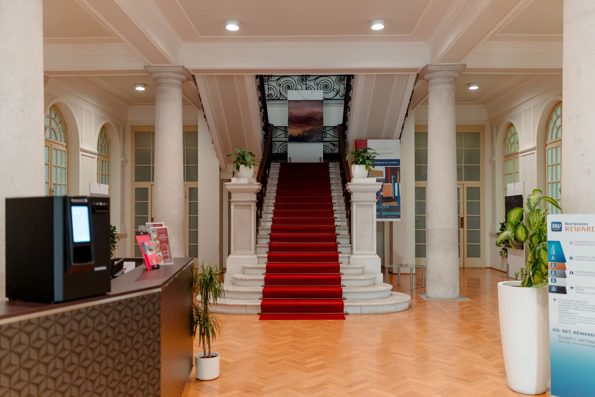 Atrio di un maestoso edificio con scalinata rivestita di moquette rossa, colonne bianche e pavimenti lucidi e luminosi.