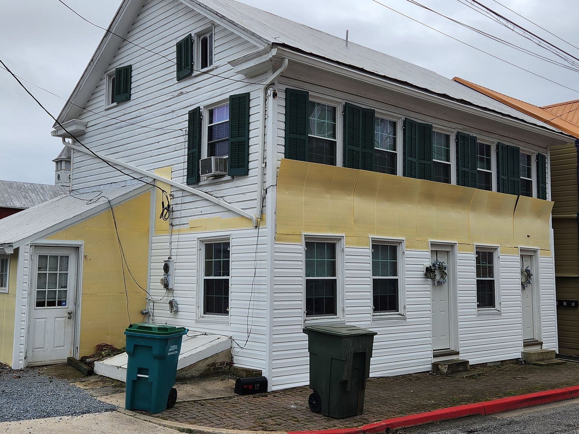 A yellow and white house with green shutters and a green trash can in front of it.