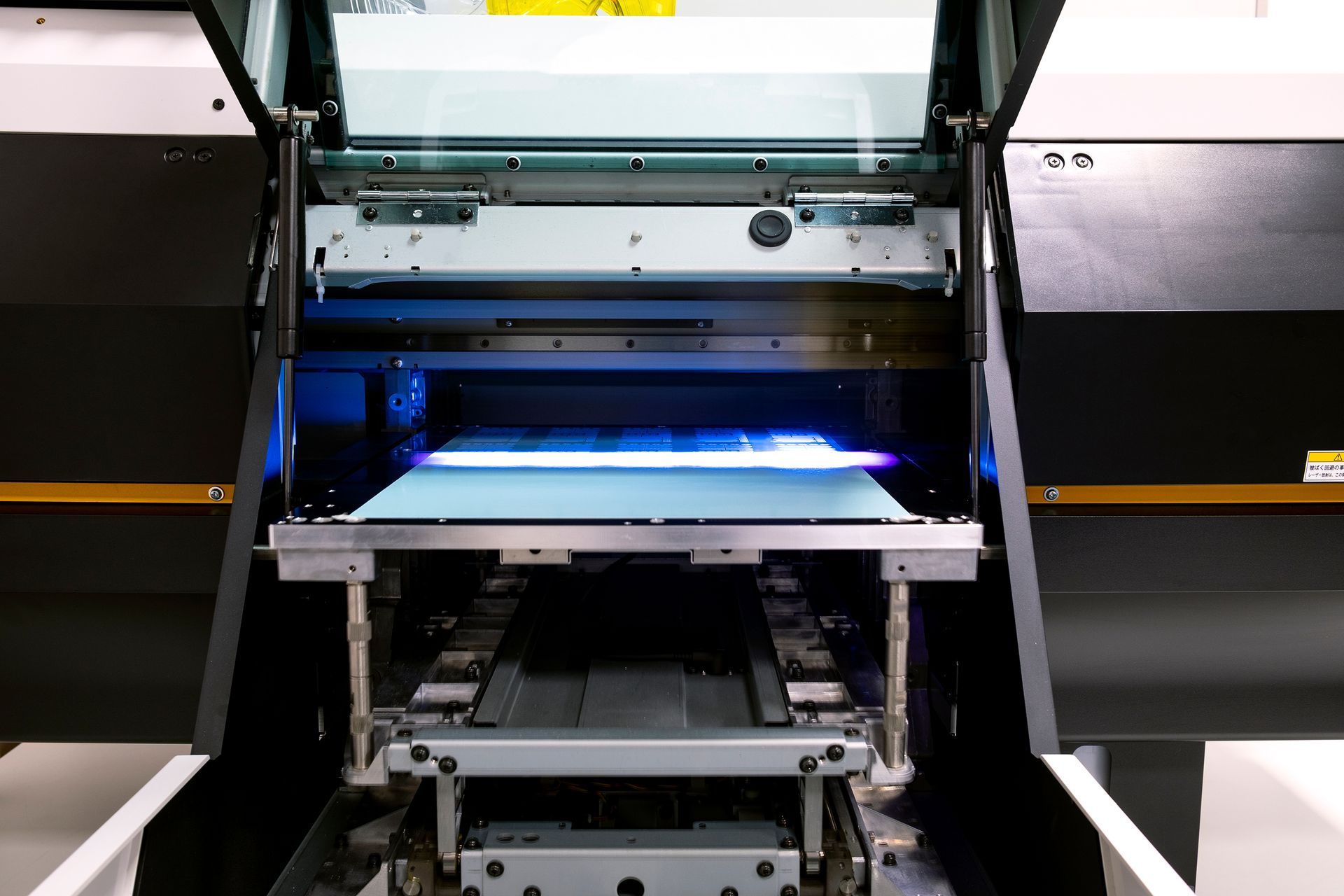 A flatbed UV printer with its lid open, showing a blue light illuminating a printing bed.