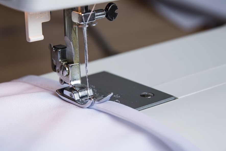 A sewing machine presser foot stitching a straight line along the edge of white fabric.