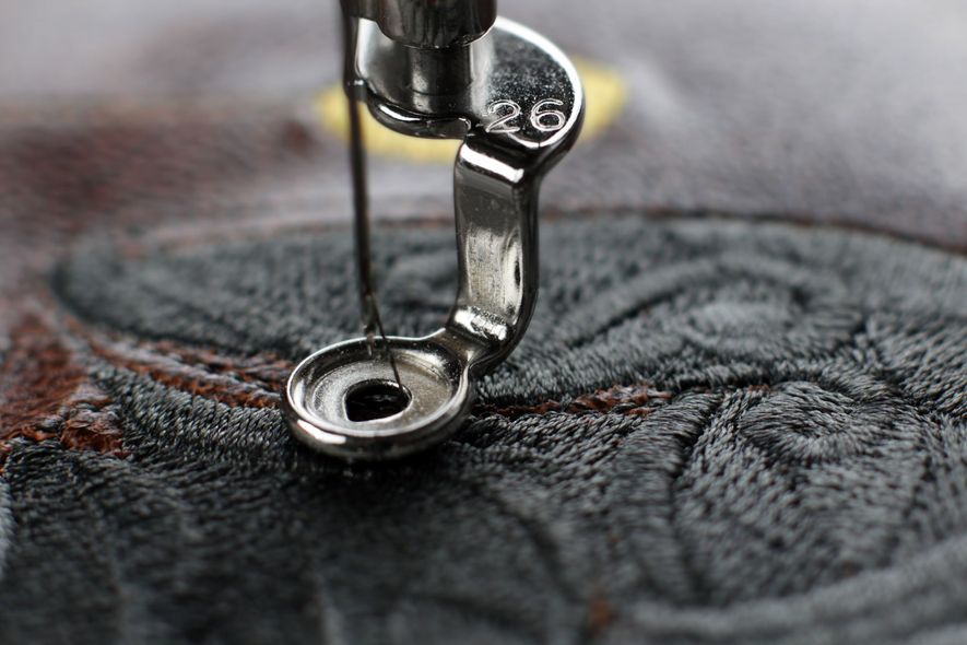 A sewing machine darning foot in action, stitching dark, detailed embroidery onto fabric.