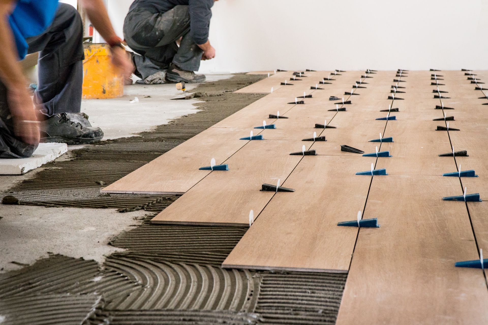 Workers installing wood flooring with blue spacers on adhesive mortar in a room