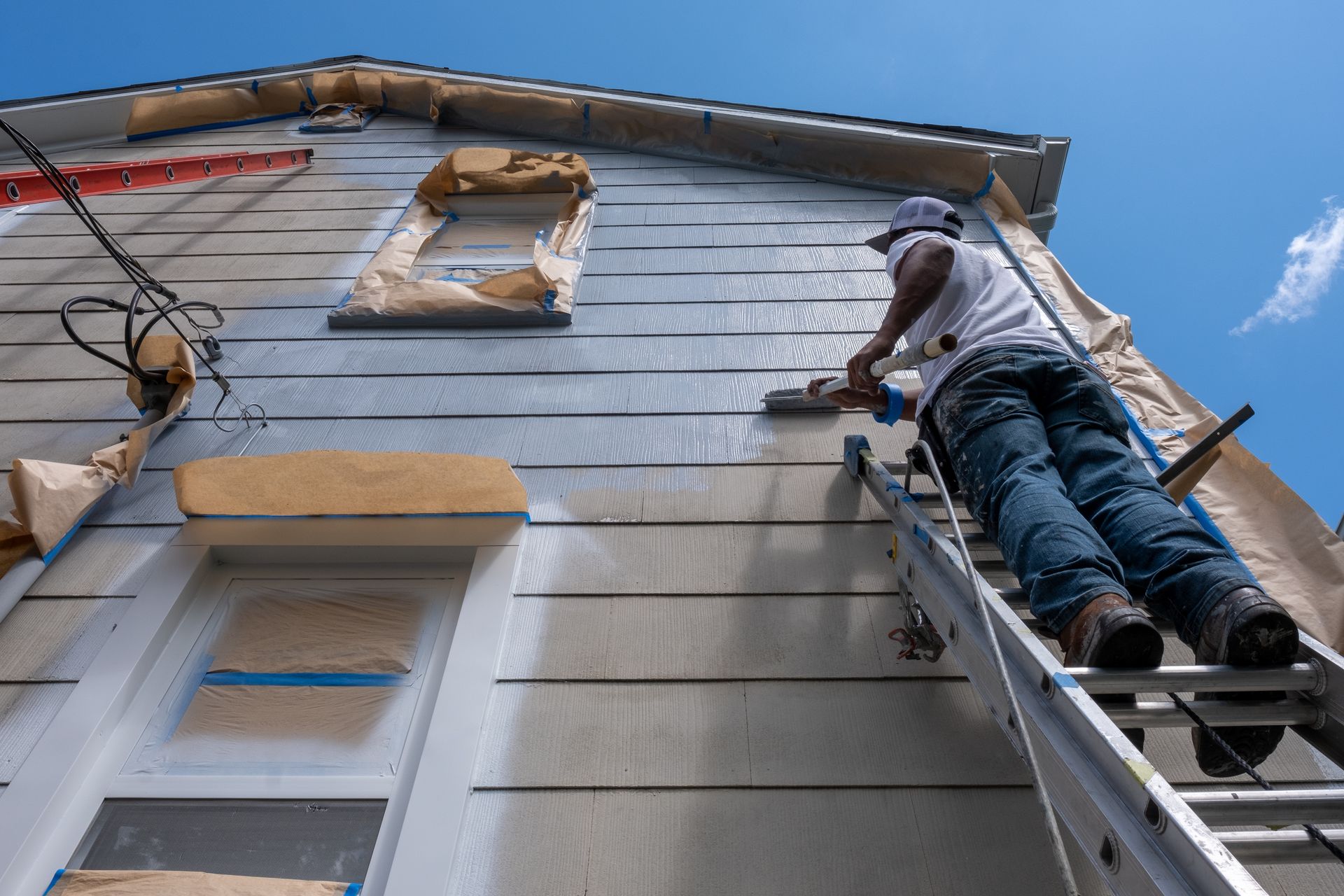 Worker on a ladder painting a house exterior beneath a blue sky