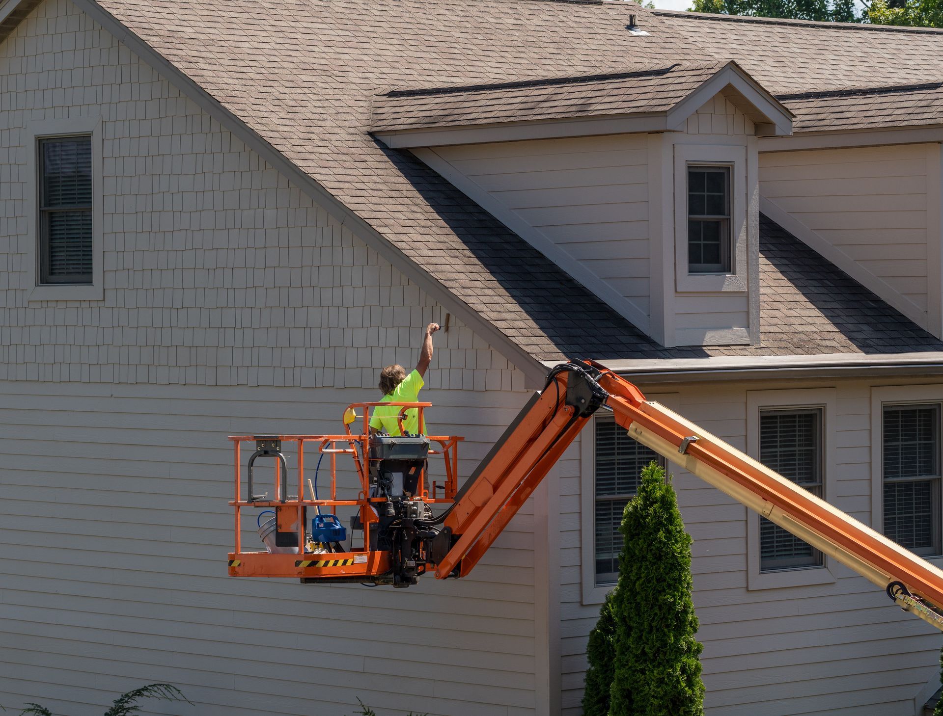 Worker in neon vest on an orange lift repairing a house roof beside dormer windows.