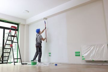 Person painting a white wall in a bright room, with a ladder and paint supplies nearby