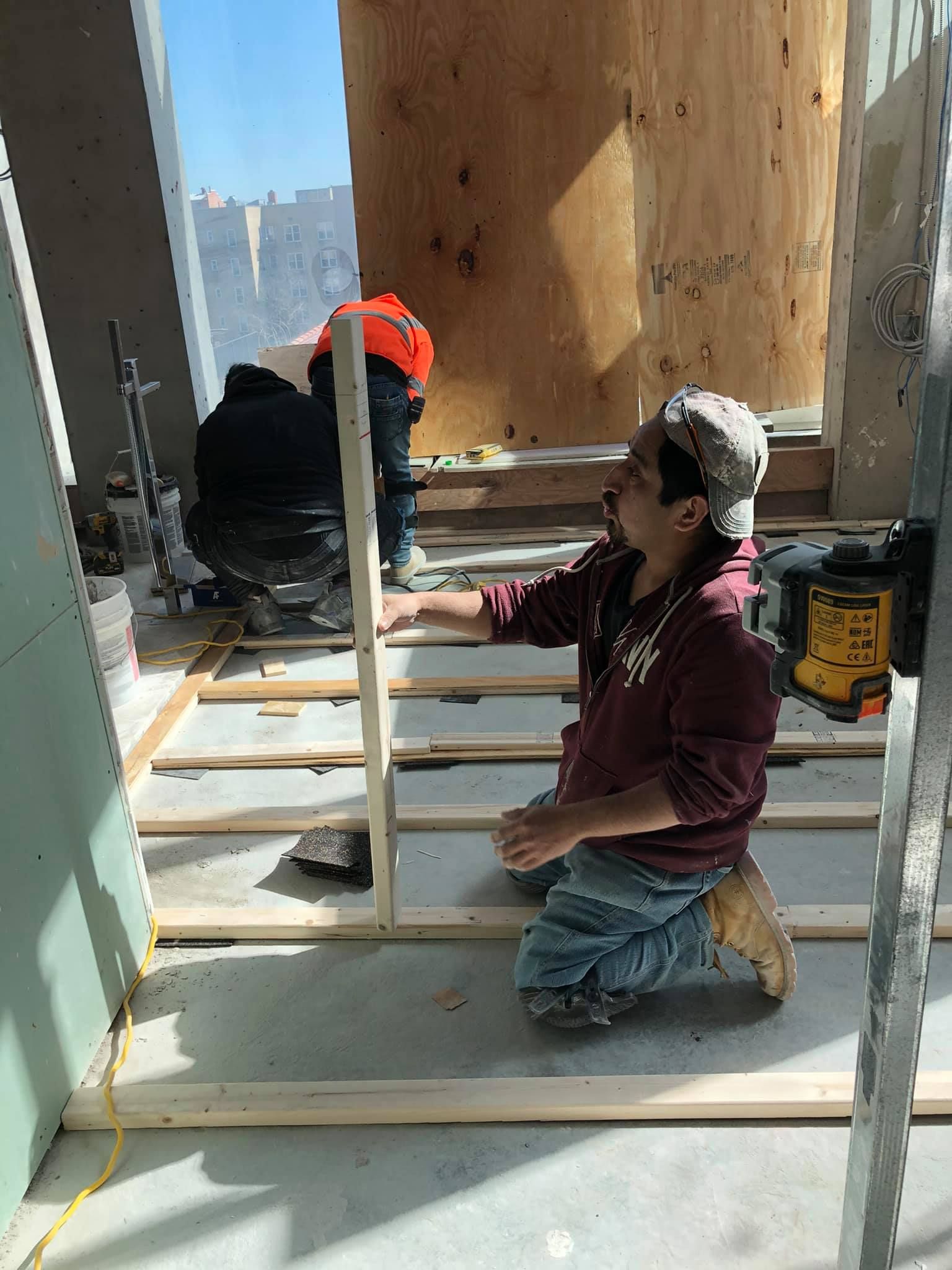 Construction workers build a wooden frame. One kneels, pointing. Others work in the background. Bright sunlight.