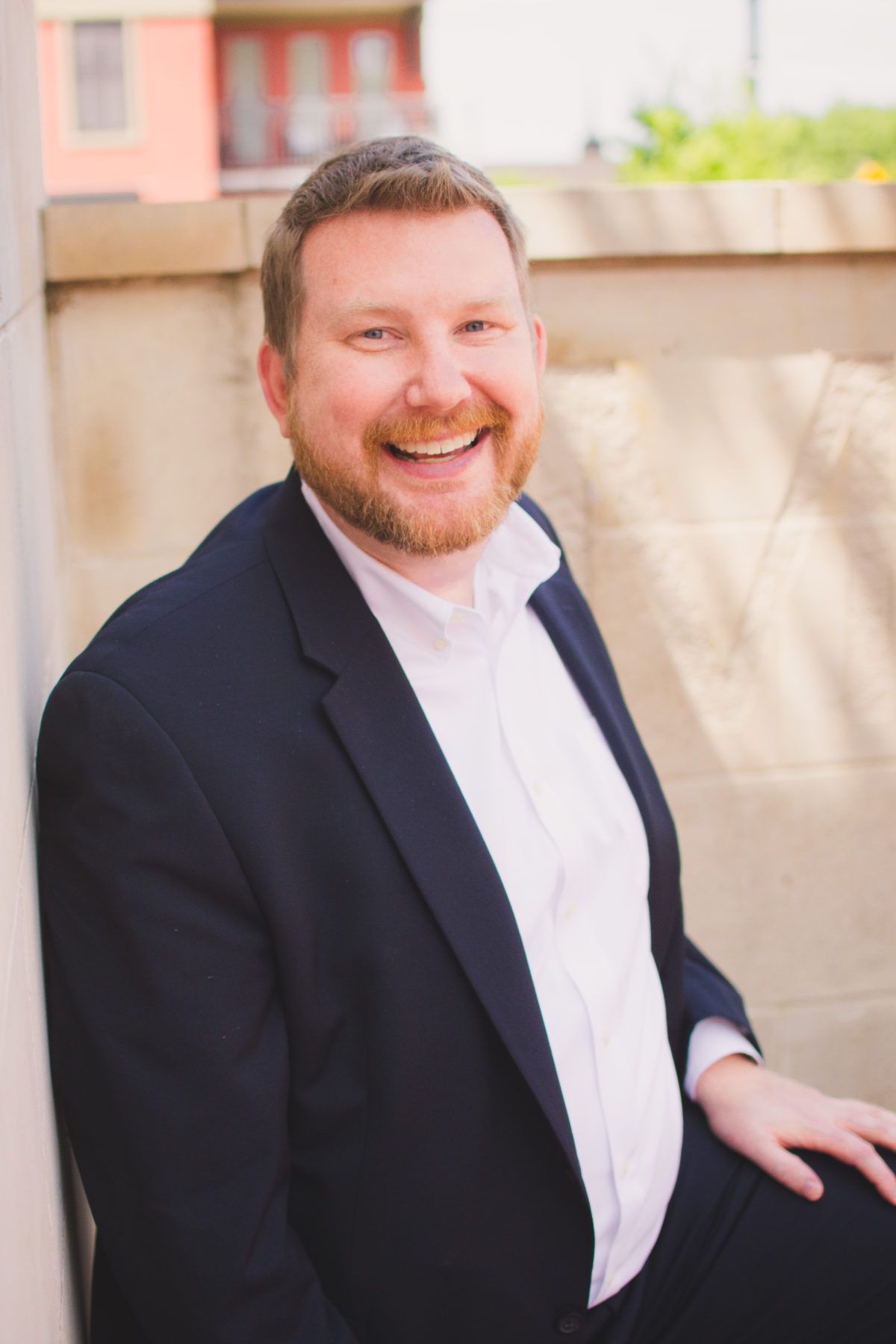 A person with a beard, wearing a navy blazer and white collared shirt, smiles while leaning against a stone wall outdoors.