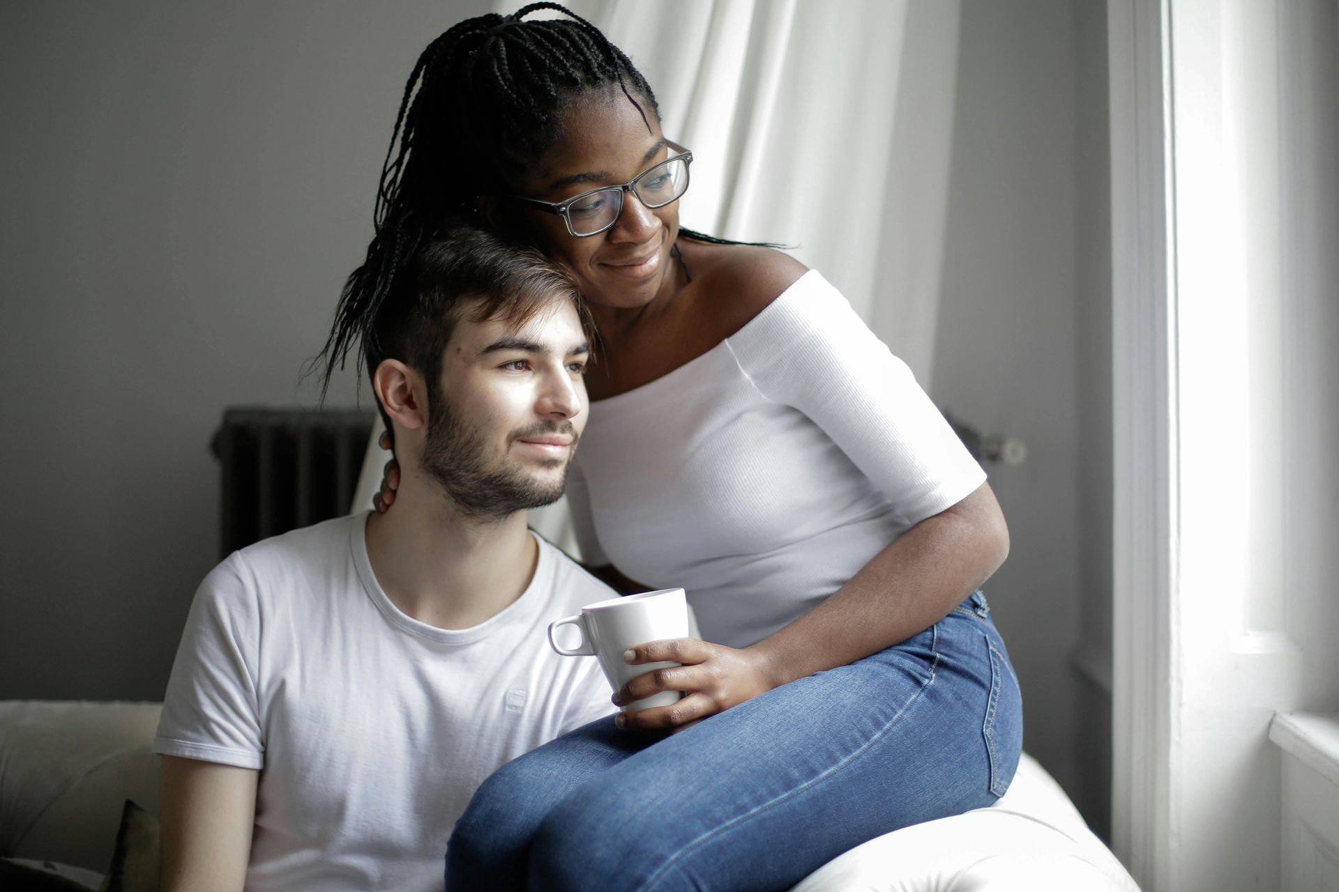 Two people sit closely by a bright window; one holds a white mug while looking outward with a peaceful expression.