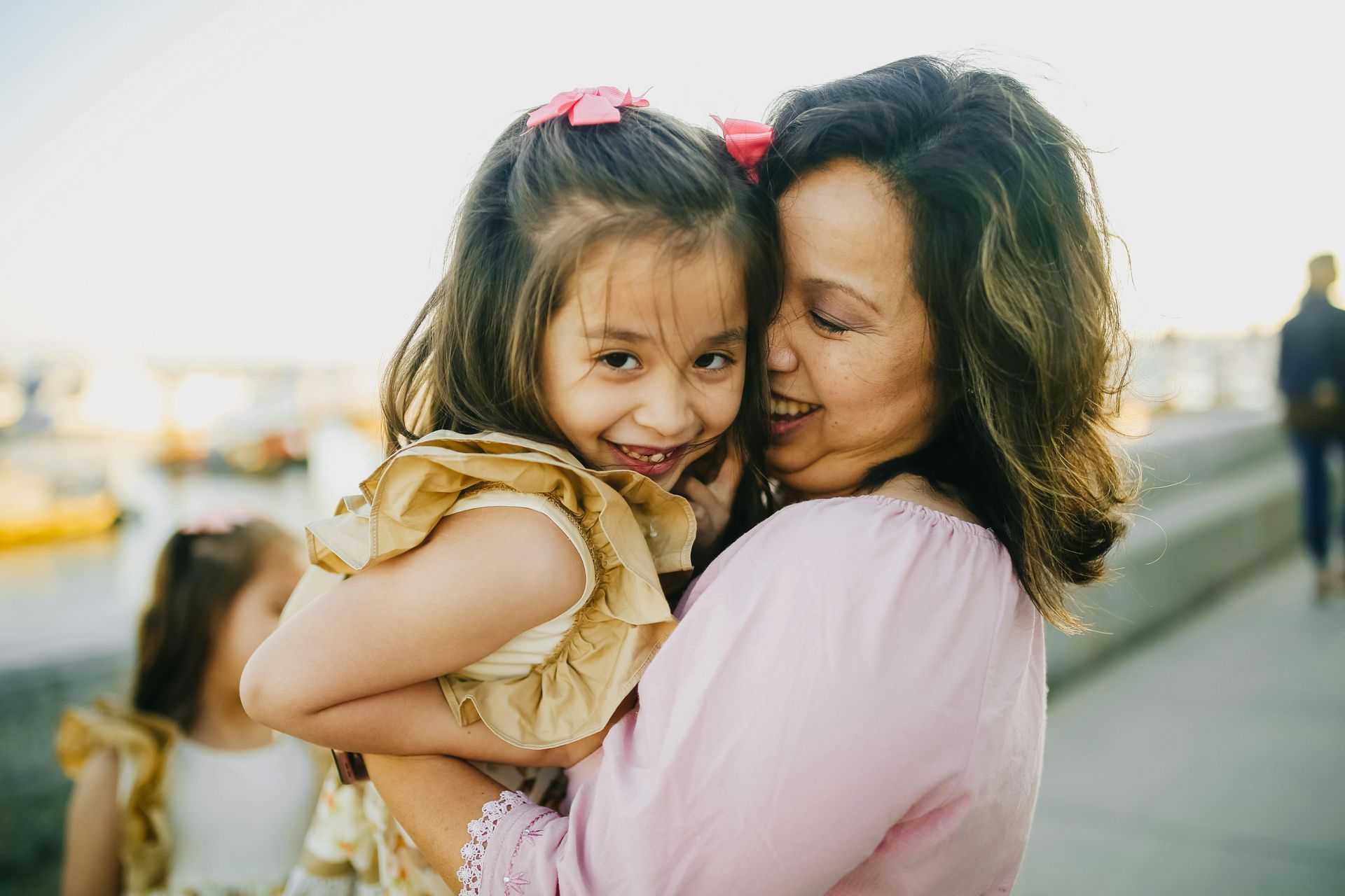 A woman in a light purple top holds a smiling child with pink hair bows in her arms, with a blurry outdoor background.