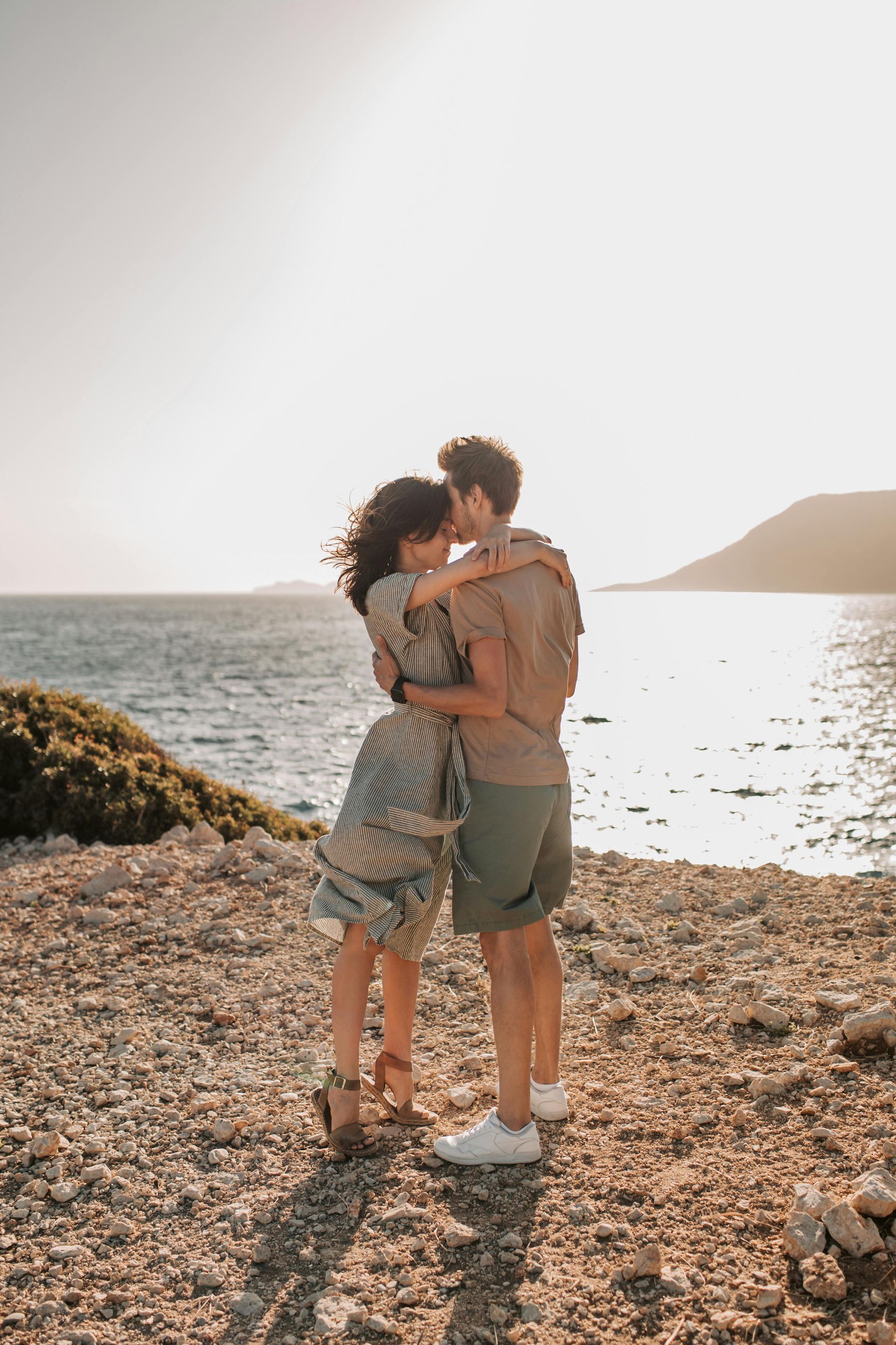 A couple embraces on a rocky shoreline overlooking the sea at sunset.