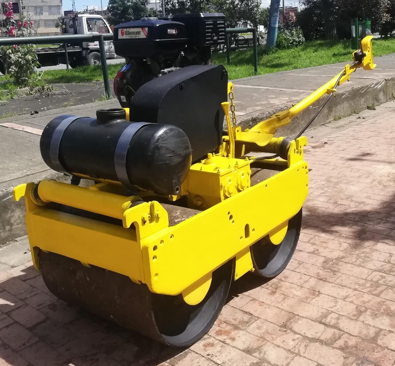 Apisonadora amarilla sobre un camino de ladrillos, con motor y depósito negros, aparcada al aire libre cerca de zonas verdes.