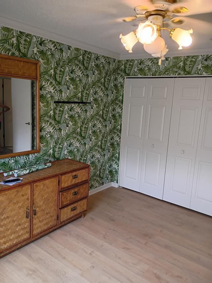 Empty bedroom with green patterned wallpaper, white closet doors, wood dresser, mirror, and ceiling fan light.