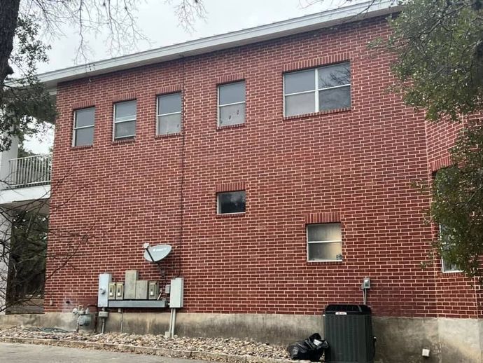 Red brick building exterior with windows, utility meters, and a satellite dish beside a wooded area