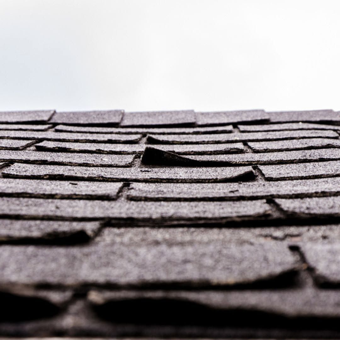 Close-up of damaged roof shingles with a lifted section against a white sky.