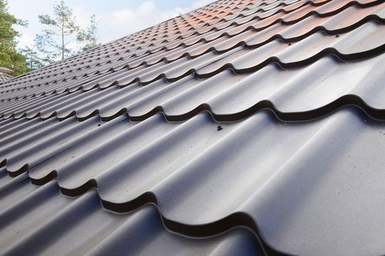 Close-up of a dark gray tiled roof with wavy metal panels and trees in the background
