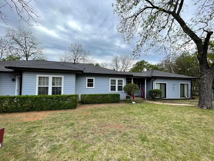 Single-story light blue house with black roof, hedges, and a large front lawn under cloudy skies