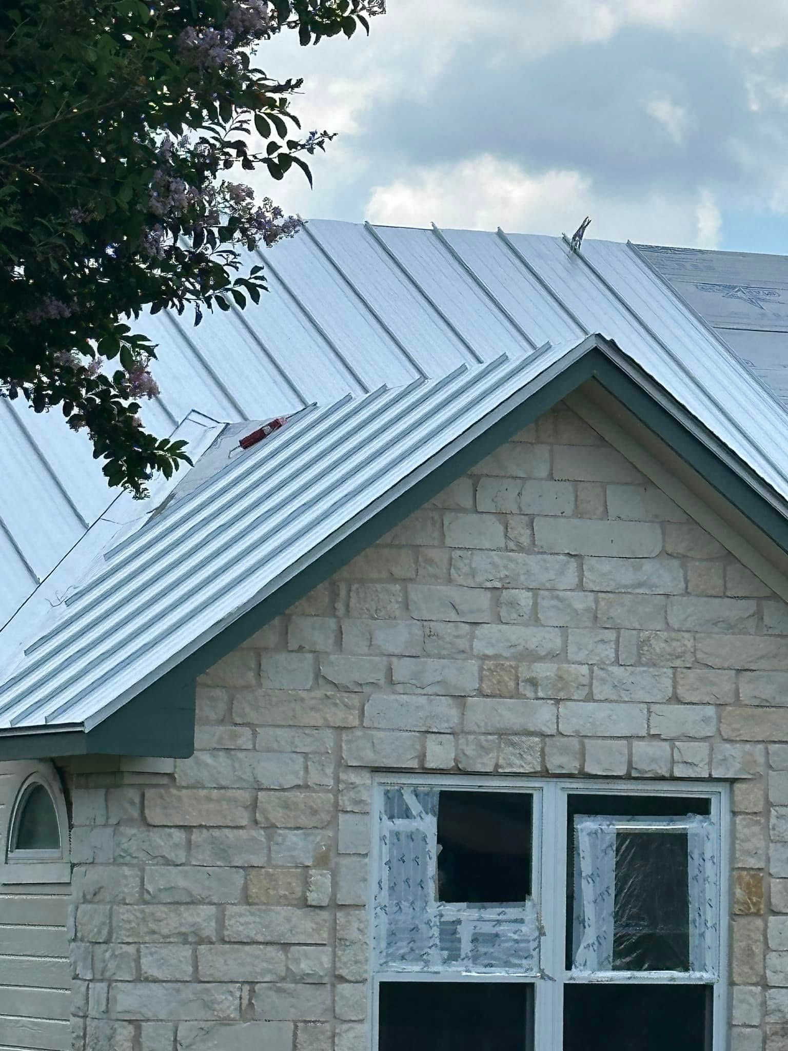 Stone house with white metal roof and broken second-floor windows under a cloudy sky.