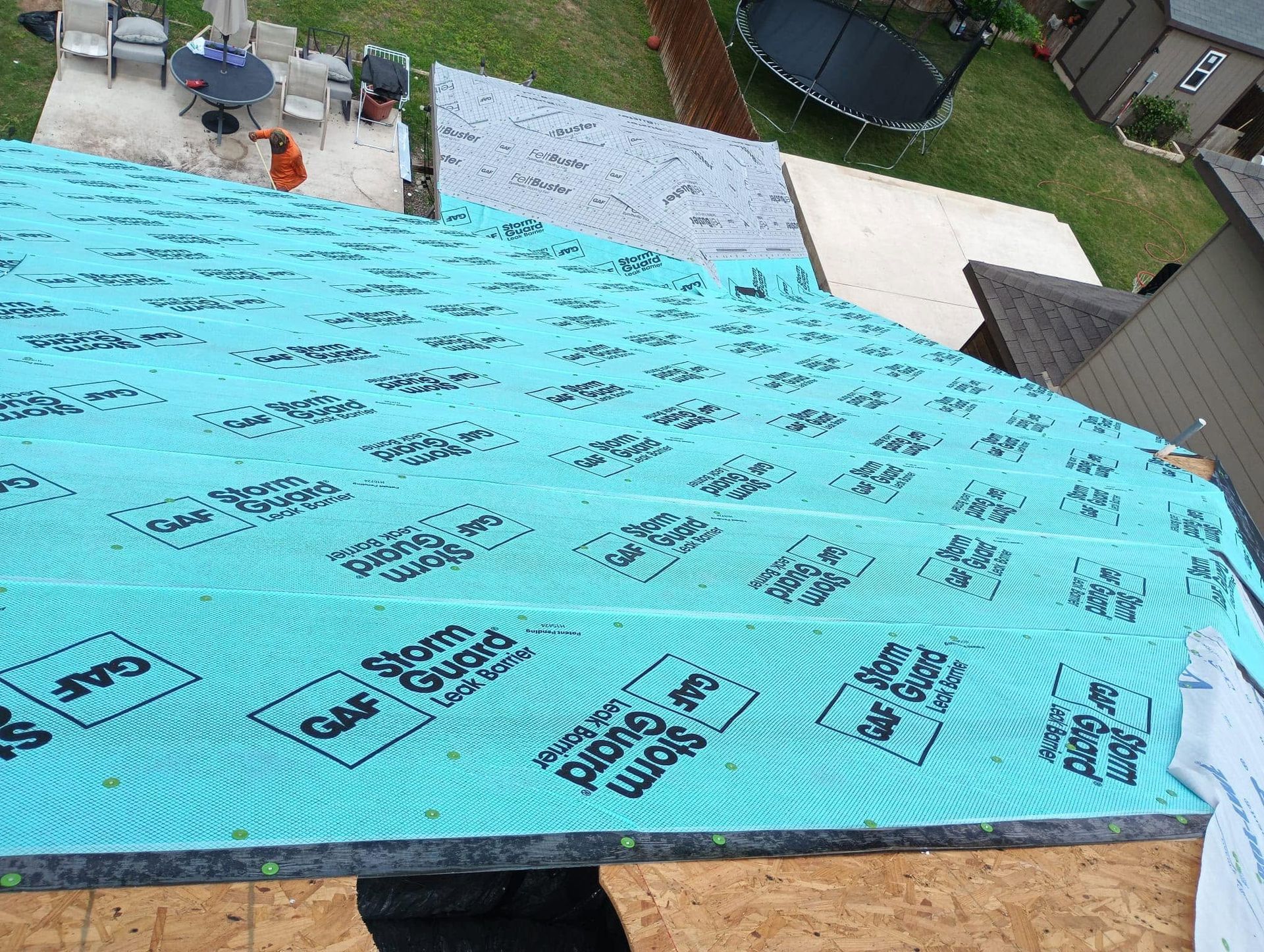 Blue roofing underlayment being installed on a roof, with workers and tools visible near the top edge.