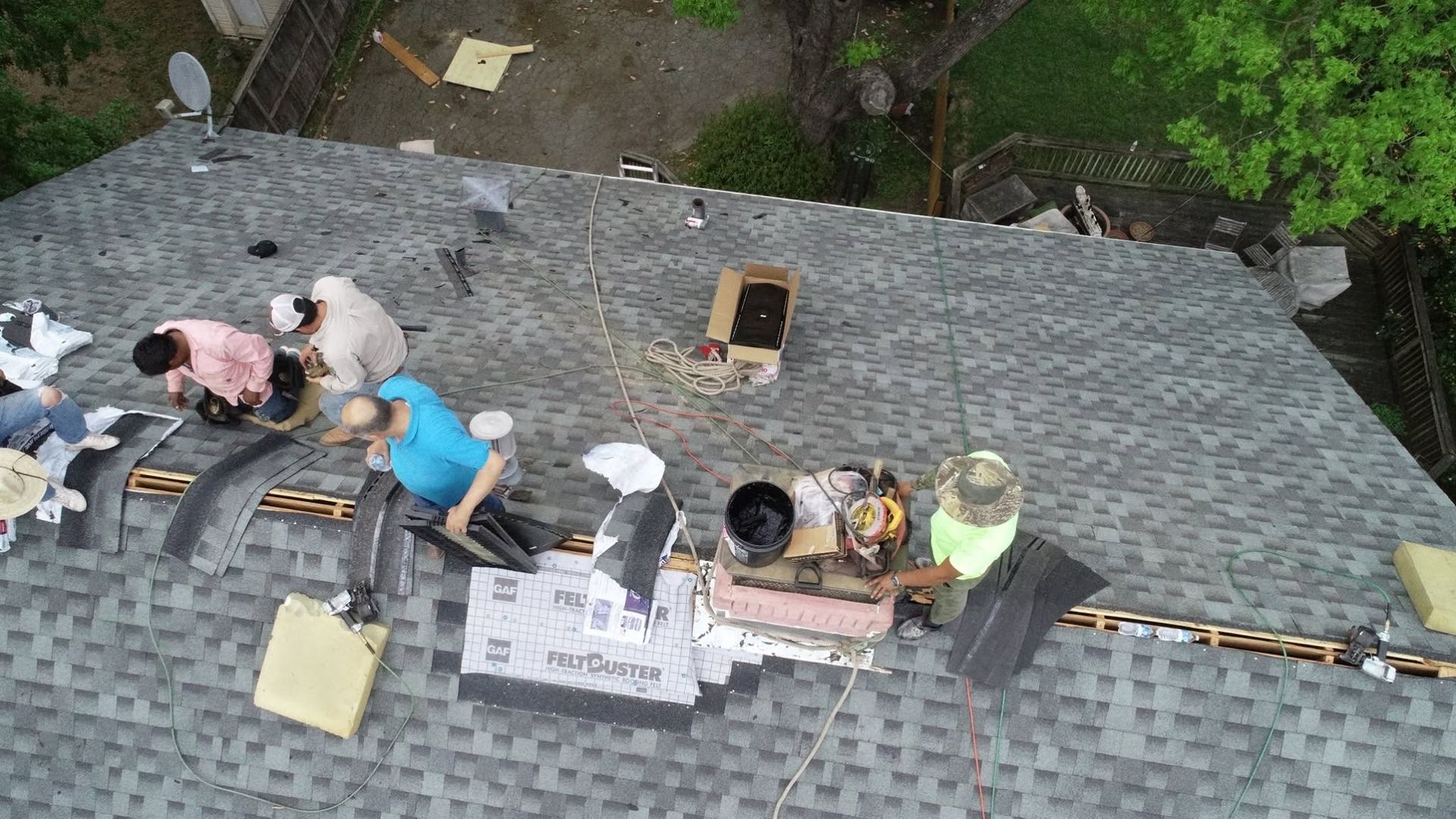 Workers on a gray shingled roof repairing equipment near an open hatch and tools.
