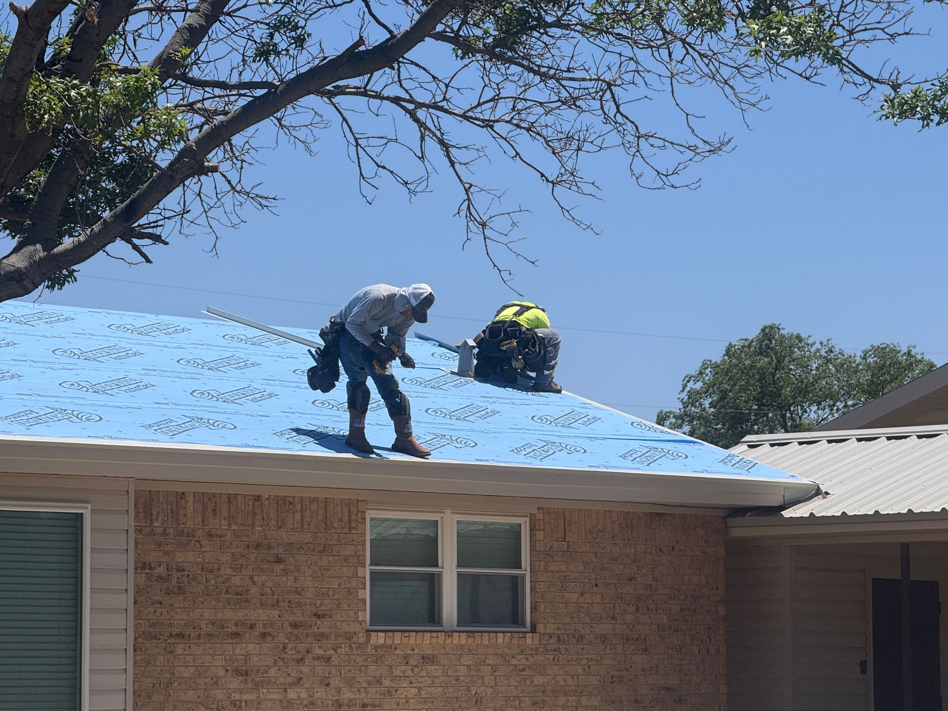 Two workers on a blue roof clearing snow above a brick house