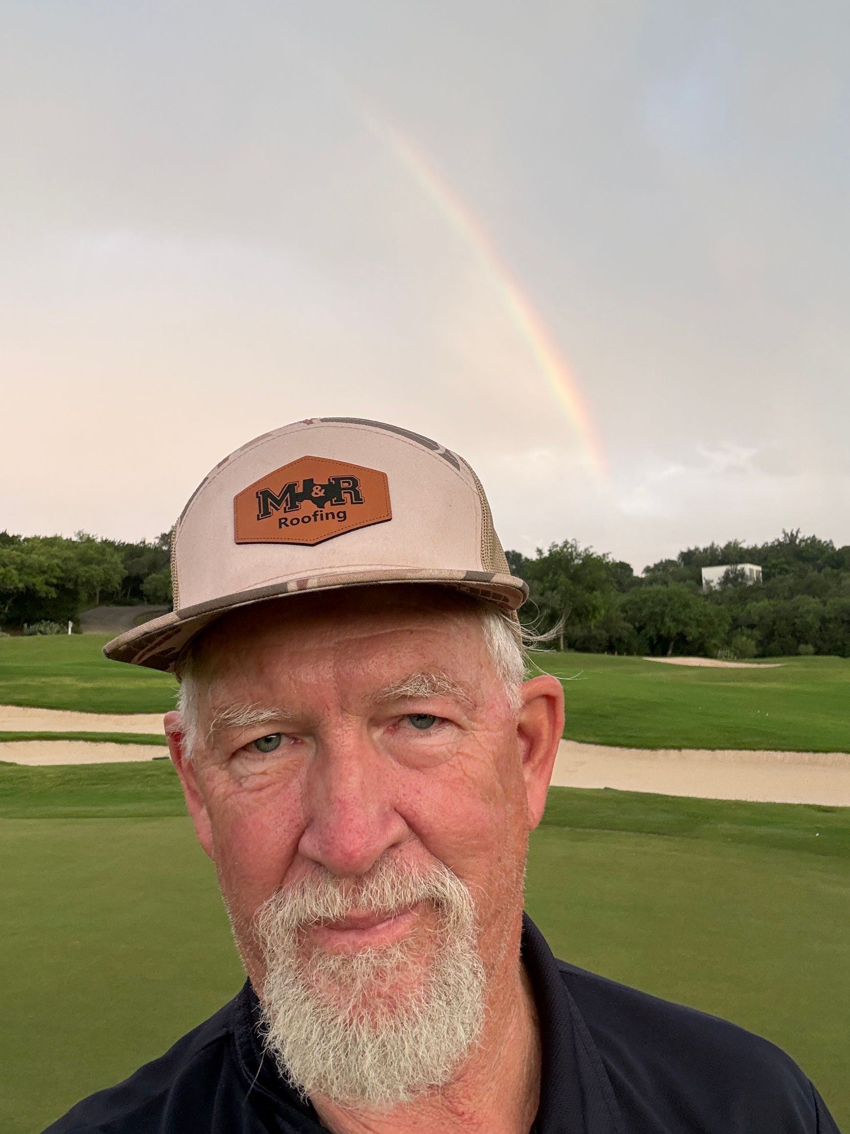 Man in a tan cap and dark shirt at a golf course with a faint rainbow in the sky