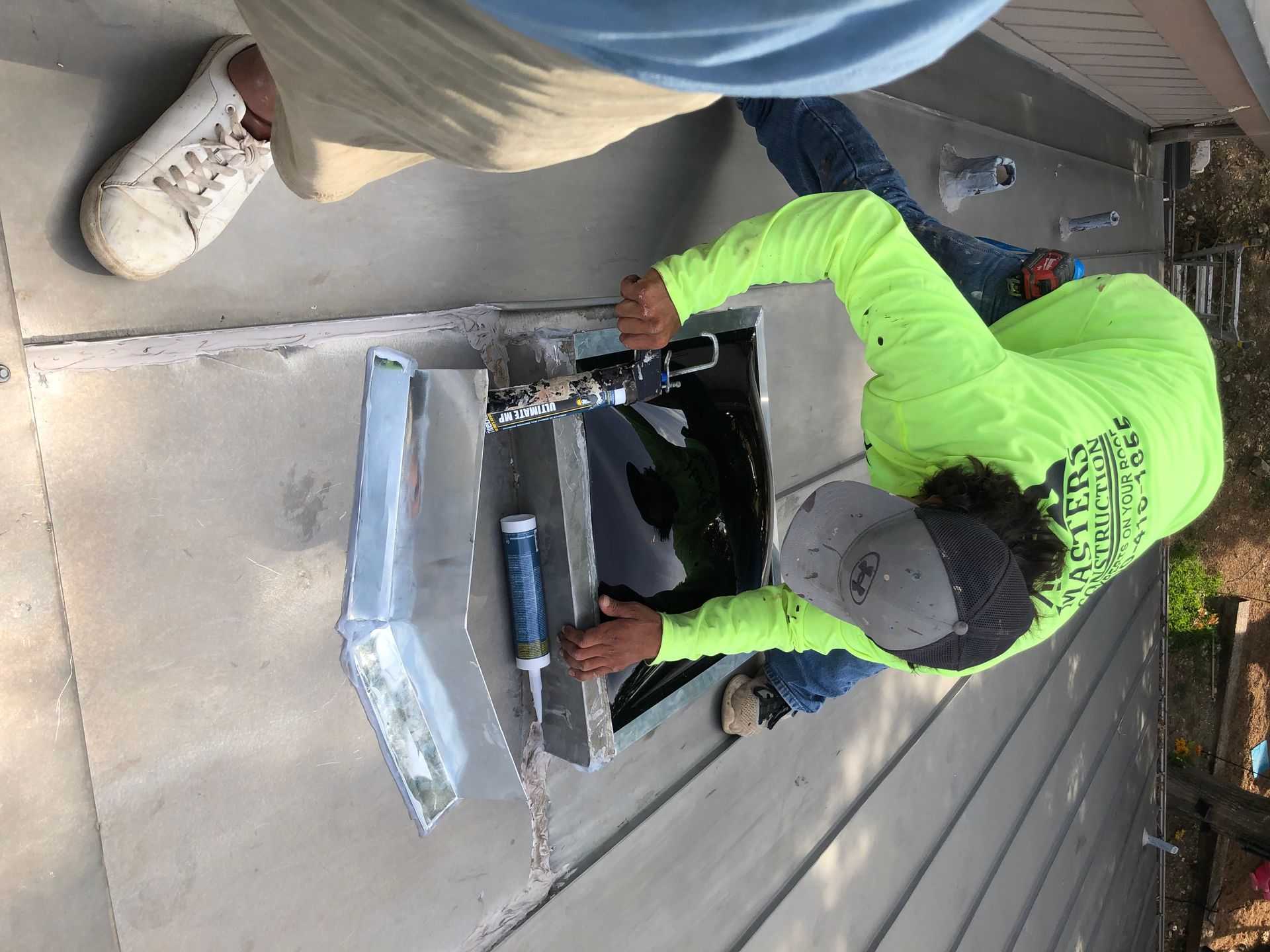 Person in neon green hoodie using a tool to work on a black rooftop vent beside metal flashing