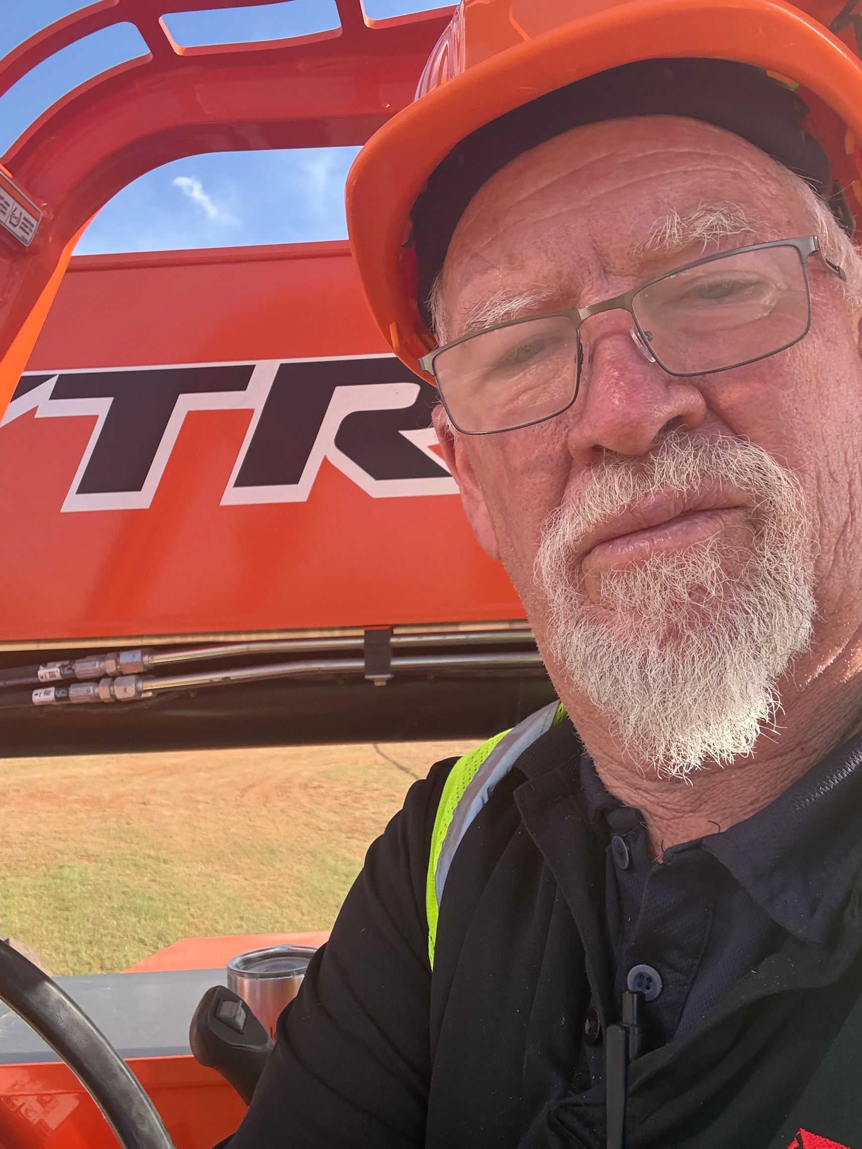 Man in glasses and orange cap beside an orange tractor, taking a selfie outdoors.