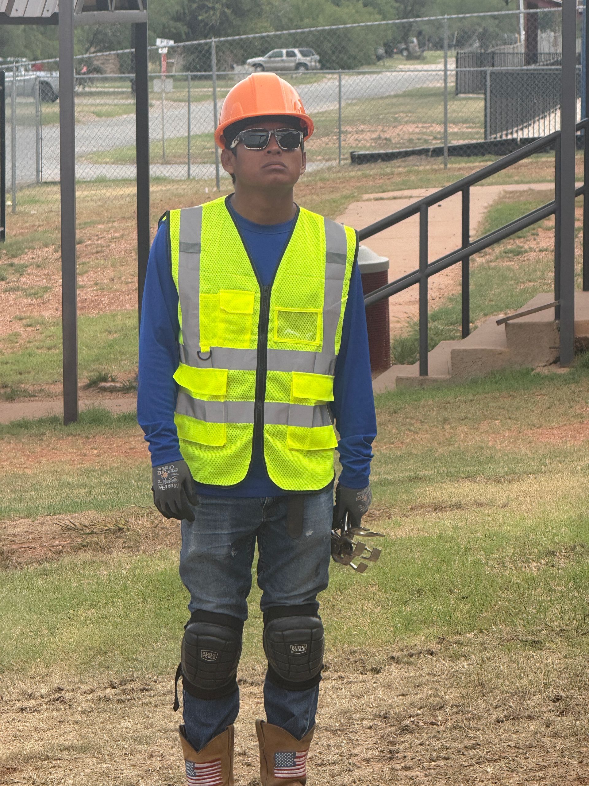 Worker in orange hard hat and yellow safety vest standing outdoors near a fence and stairs.