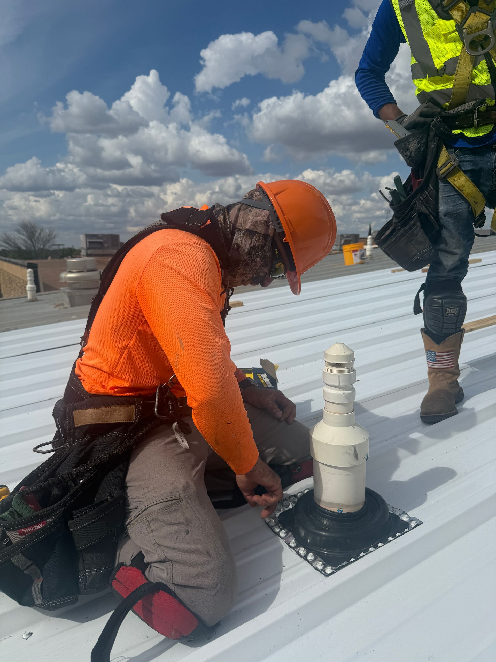 Worker in orange safety gear kneels on a rooftop beside a vent pipe while another worker stands nearby.