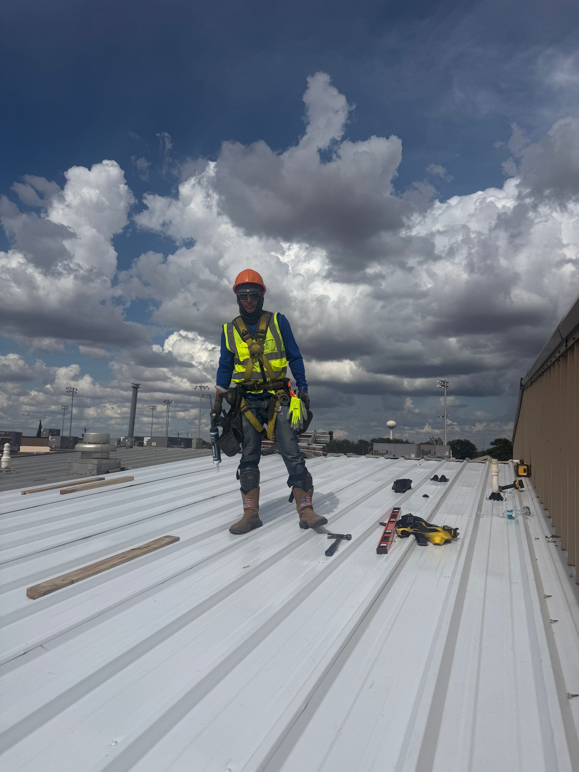 Worker in safety gear standing on a white rooftop under a cloudy sky, with tools nearby.