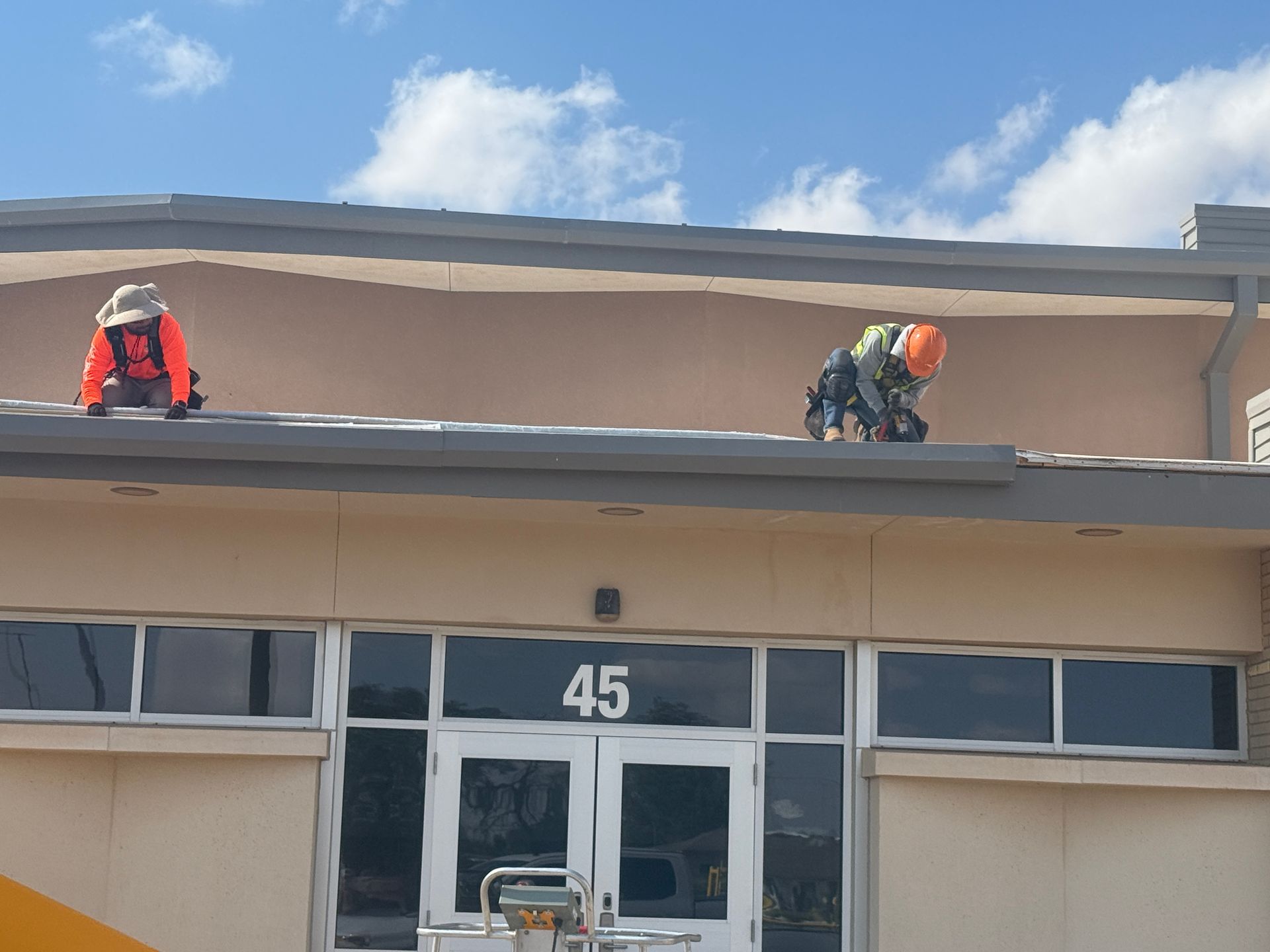 Two workers on a rooftop above a building entrance, one in red and one in orange safety gear.