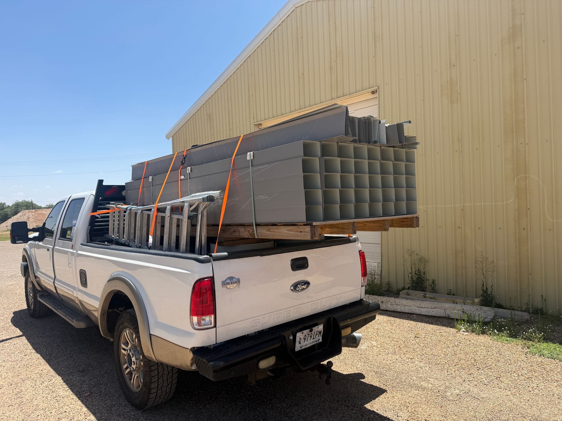 White pickup truck loaded with stacked metal pipes beside a tan warehouse on a gravel lot
