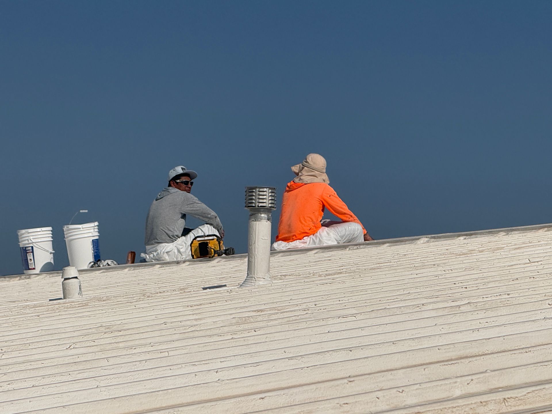 Two workers sit on a white rooftop under a clear blue sky, one in orange jacket and one in gray.