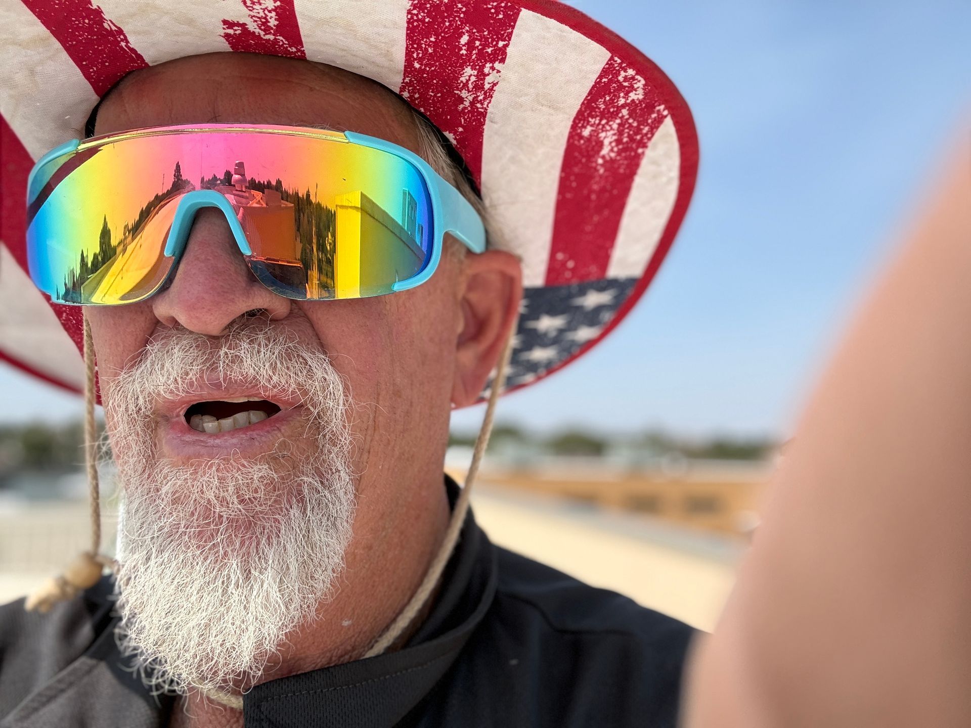 Man wearing reflective sunglasses and a red-and-white striped hat at the beach, with a white beard and black shirt