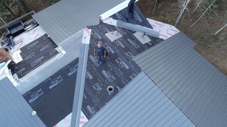 Roofer working on a partially covered roof with gray shingles and black underlayment, viewed from above.