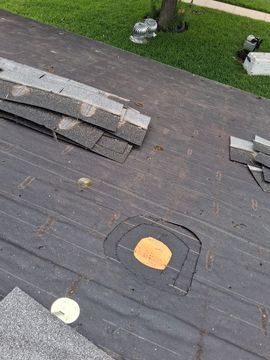 Rooftop with stacked shingles, a vent flashing, and a grassy yard visible beyond the roof edge.