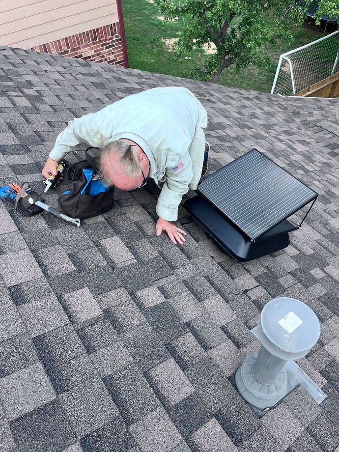 Worker kneeling on a shingled roof beside a black vent cap and rooftop vent pipe.