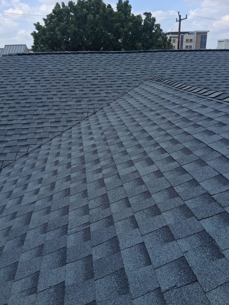 Gray shingled roof on a building, viewed from a low angle with trees and a distant structure in the background