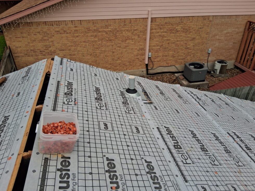Roofing underlayment covered with scattered orange fasteners beside a brick house and patio furniture