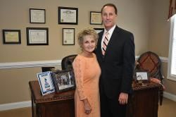 Two people posing in an office, with framed certificates and photos on the wall behind them