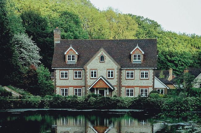 Red-roofed cottage by a calm pond, surrounded by lush green trees, with its reflection in the water.