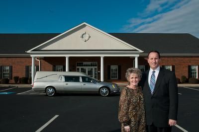 A couple stands outside a brick building with a white portico; a silver hearse is parked nearby.