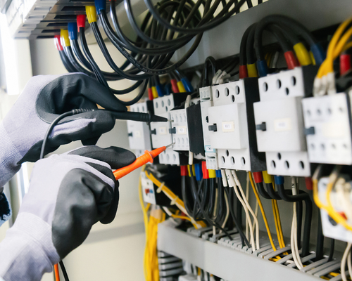 Gloved technician testing wiring in an electrical control panel with cables and circuit breakers