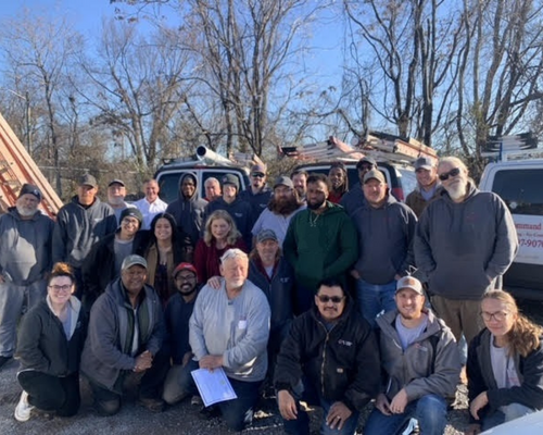 Large group posing outdoors on a sunny day in front of parked vehicles and bare trees