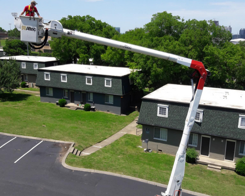 Two workers in a cherry picker repairing a rooftop on dark green apartment buildings.