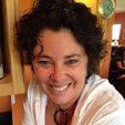 Smiling woman with dark curly hair, wearing glasses, white shirt, and reddish necklace.