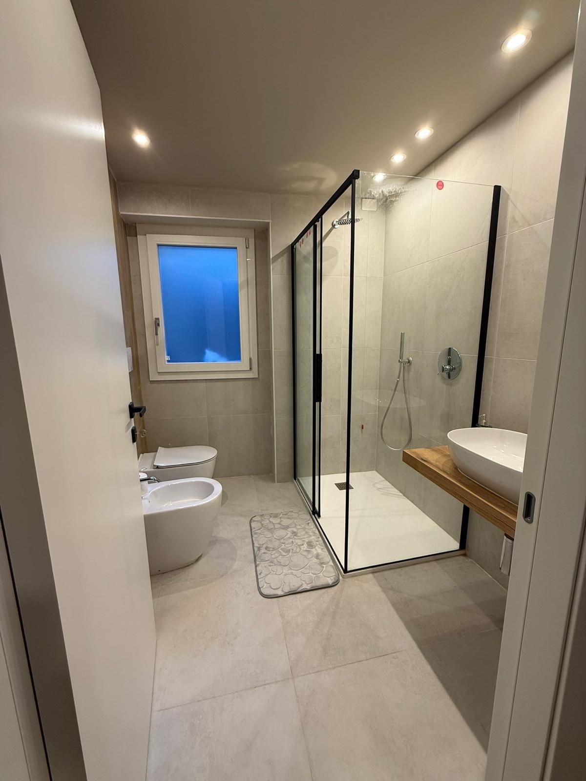 Bagno moderno con doccia in vetro, lavabo, WC e finestra illuminata di blu.