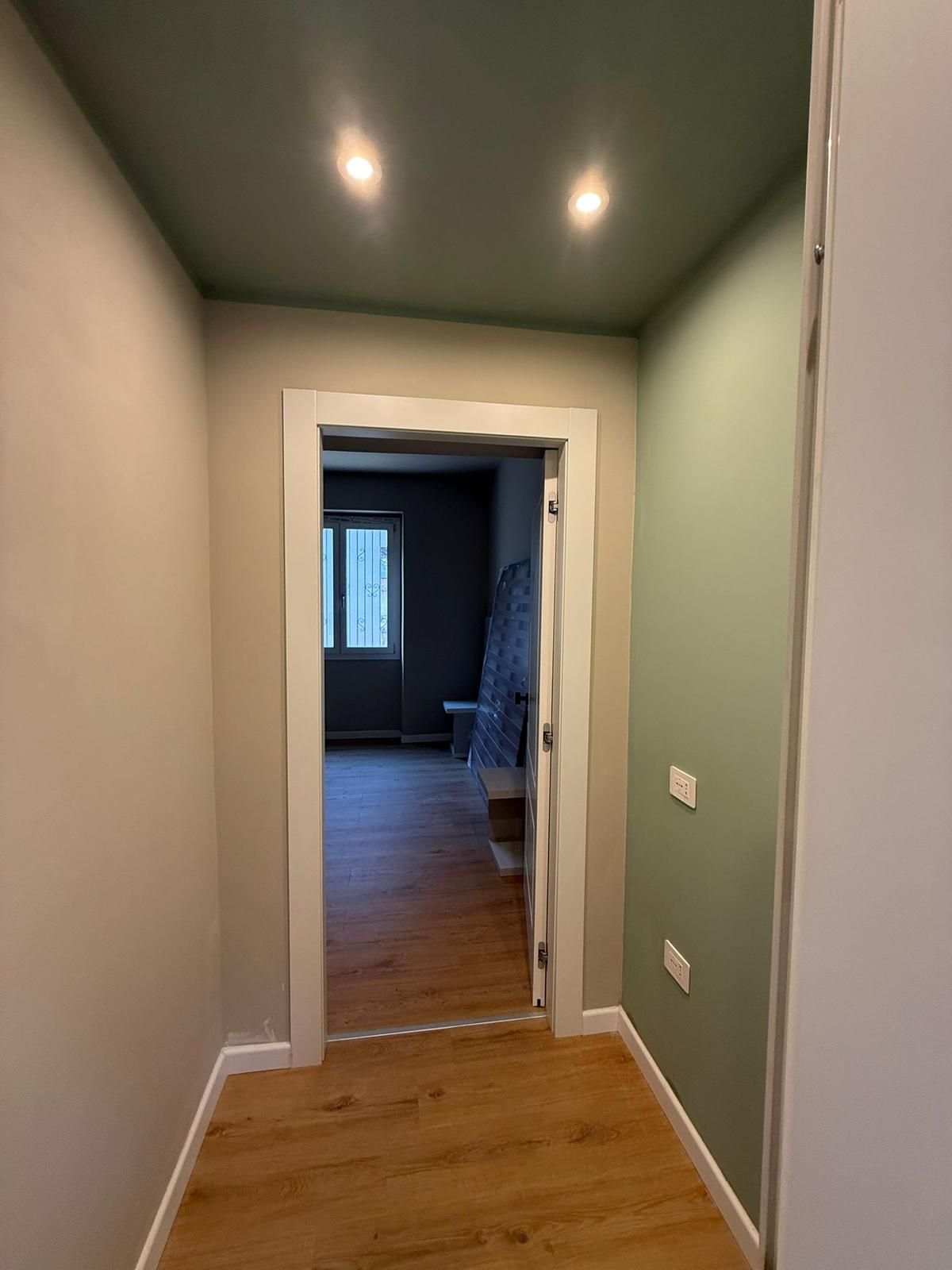 Stretto corridoio con pareti verdi, pavimento in legno e una porta che conduce a una stanza poco illuminata con una finestra.